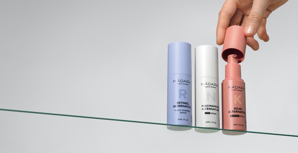 A hand removes the cap from a coral-colored serum bottle beside two other pastel bottles on a glass shelf, showcasing three facial serums from MÁDARA Organic Skincare: Retinol Alternative, Niacinamide Alternative, and Kojic Alternative, against a minimal light background