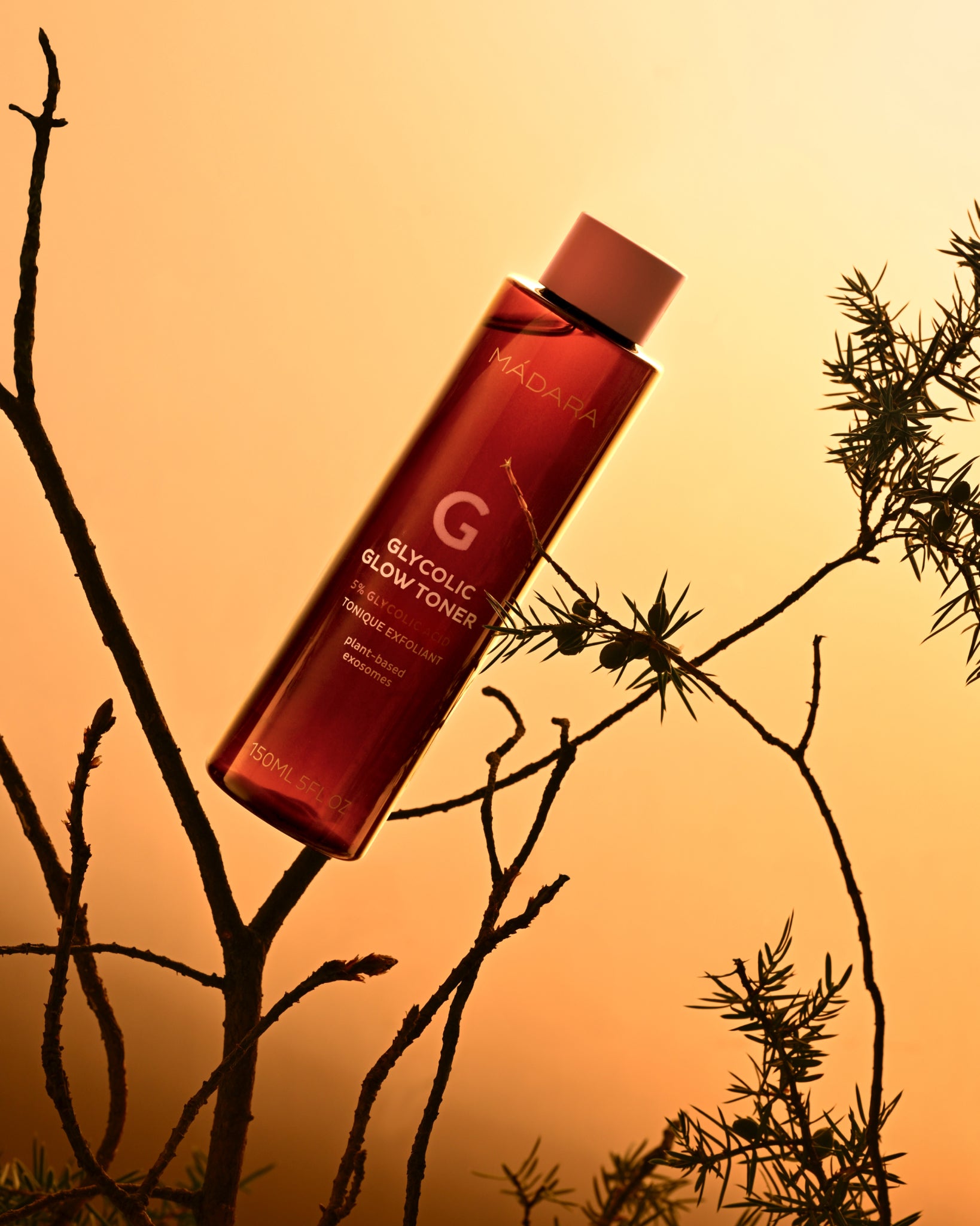 MADARA Glycolic Glow Toner bottle on a branch against an orange sunset sky
