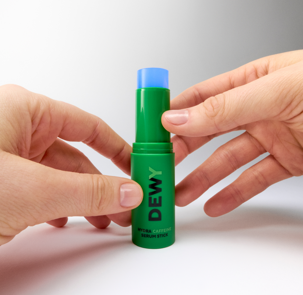 Hands holding MÁDARA DEWY Hydra Caffeine Serum Stick with the blue balm partially extended against a clean light background