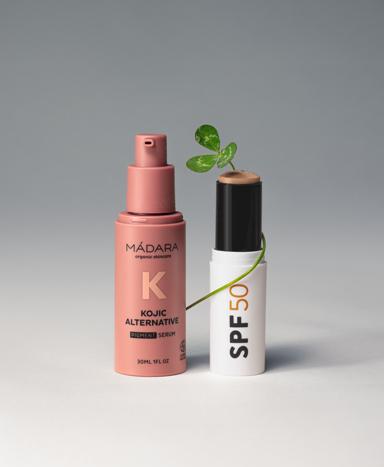 MÁDARA Kojic Alternative pigment serum and SPF50 stick displayed on a grey background with a green plant stem