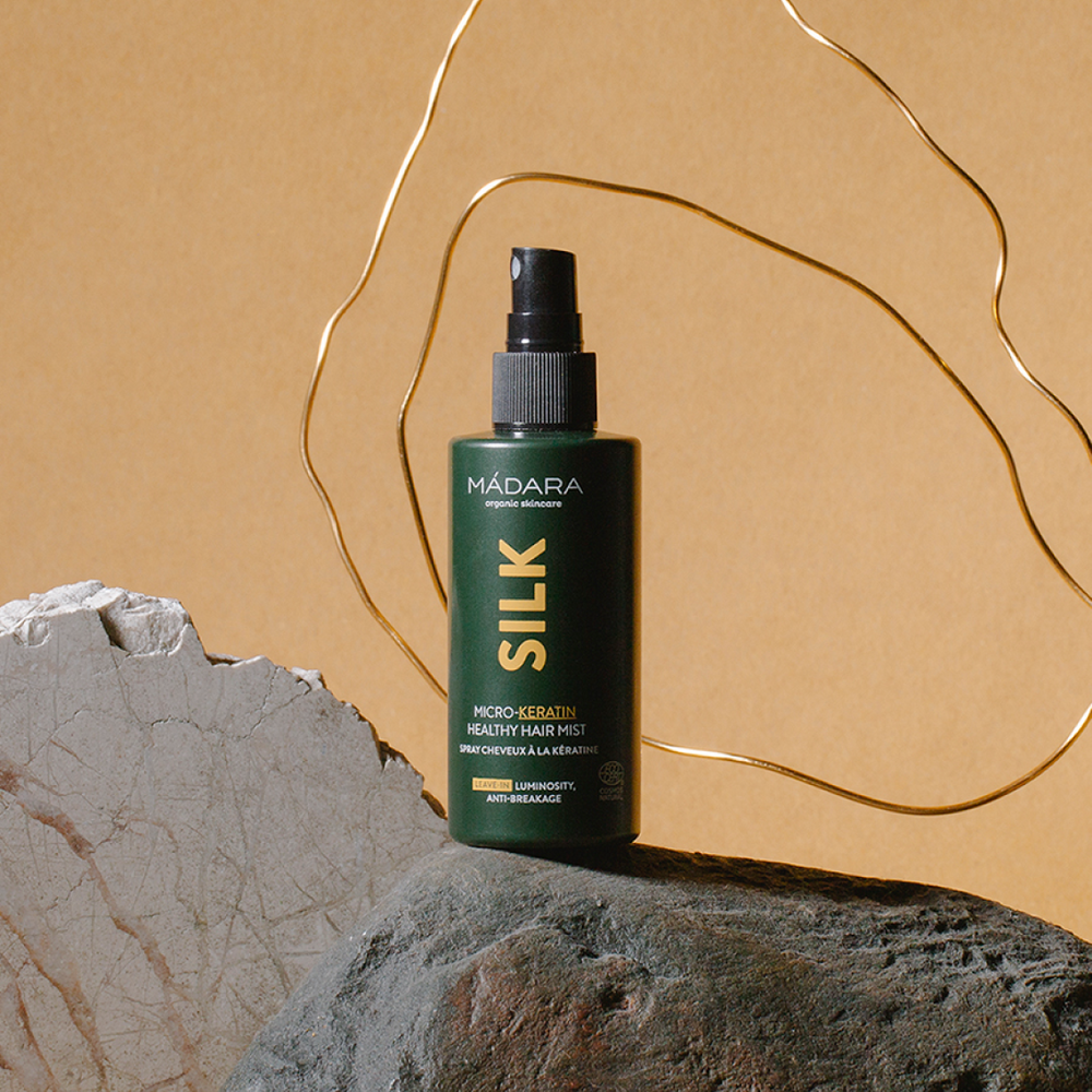 MÁDARA Silk Micro-Keratin Healthy Hair Mist bottle placed on a stone surface with abstract gold shapes in the background