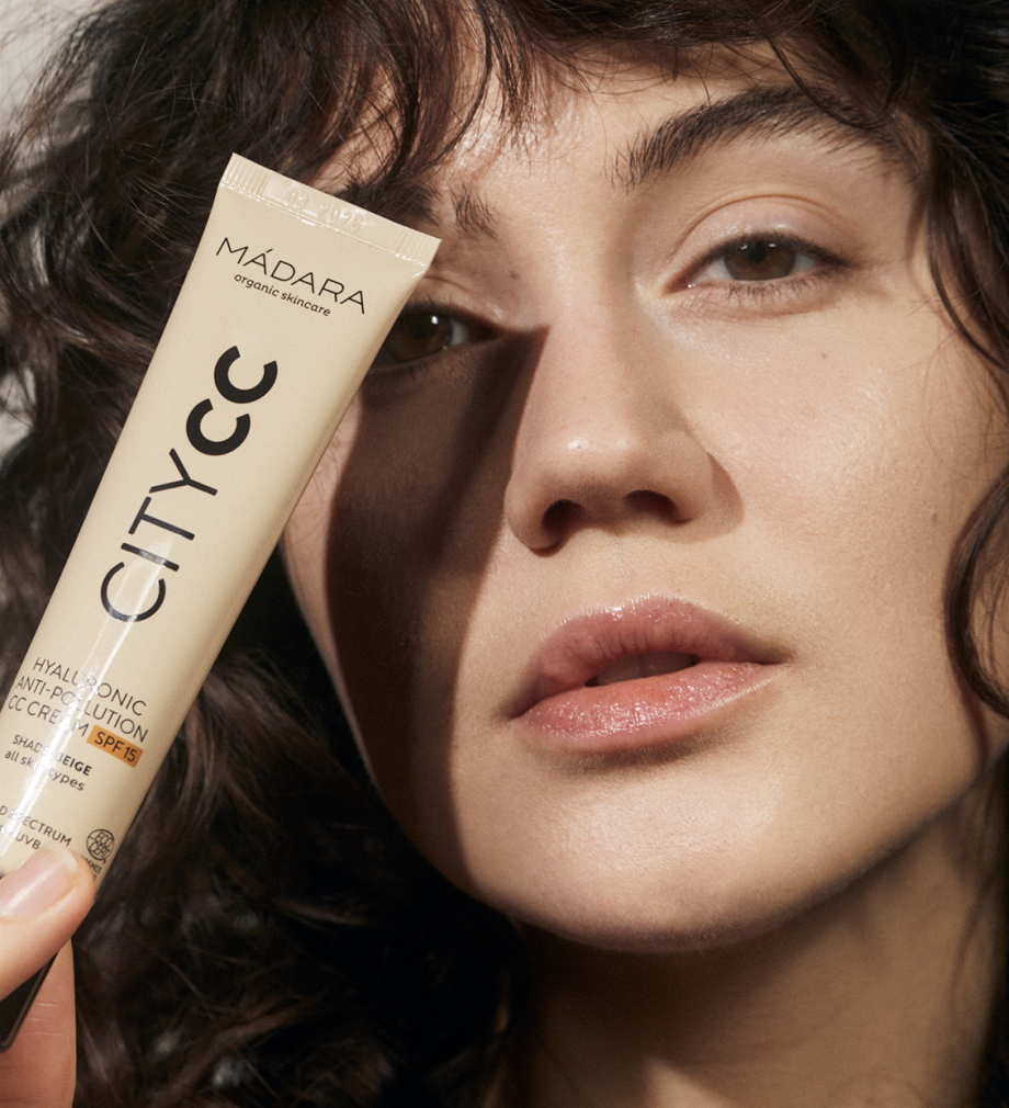 Model holding MÁDARA CITY CC Hyaluronic Anti-Pollution CC Cream SPF15 tube beside her face, showing natural glowing skin and lightweight complexion coverage