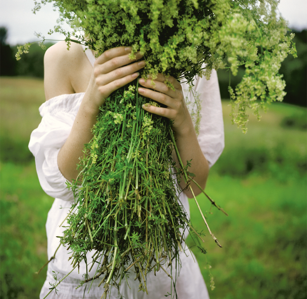 Person in a white dress holding a large bouquet of fresh green wildflowers, covering their face, in a grassy outdoor setting