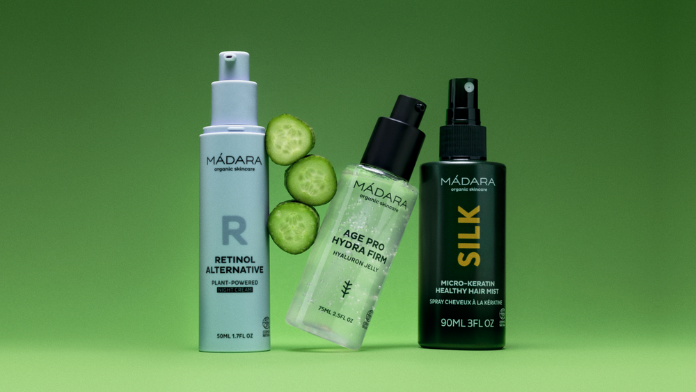 Three MÁDARA organic skincare products on a green background Retinol Alternative night cream, Age Pro Hydra Firm hyaluron jelly, and Silk Micro-Keratin Healthy Hair Mist, with sliced cucumber