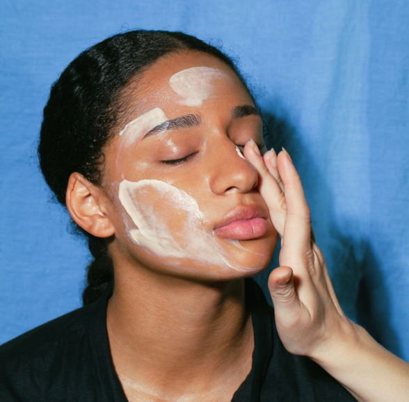 A close-up of a woman with madara cleansing balm applied in thick patches, applying it to her cheek with her hand against a solid blue background.