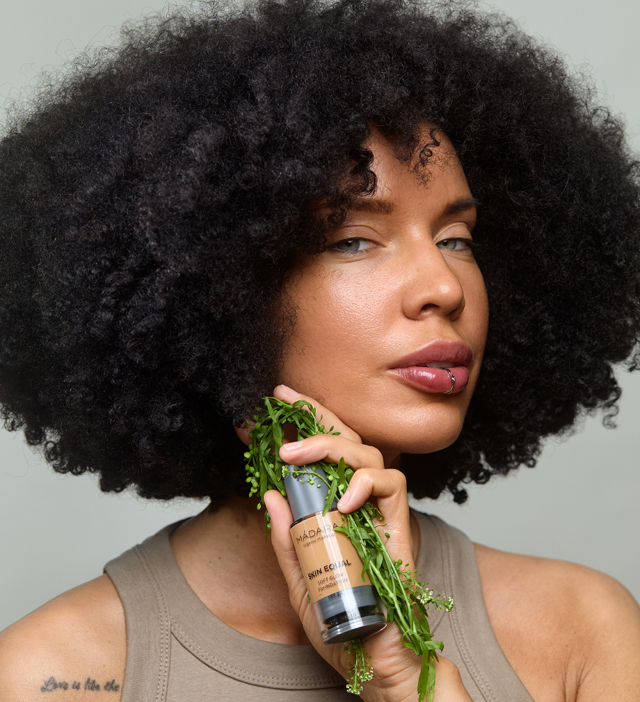 Woman holding MÁDARA Skin Equal Soft Glow Foundation surrounded by green botanicals