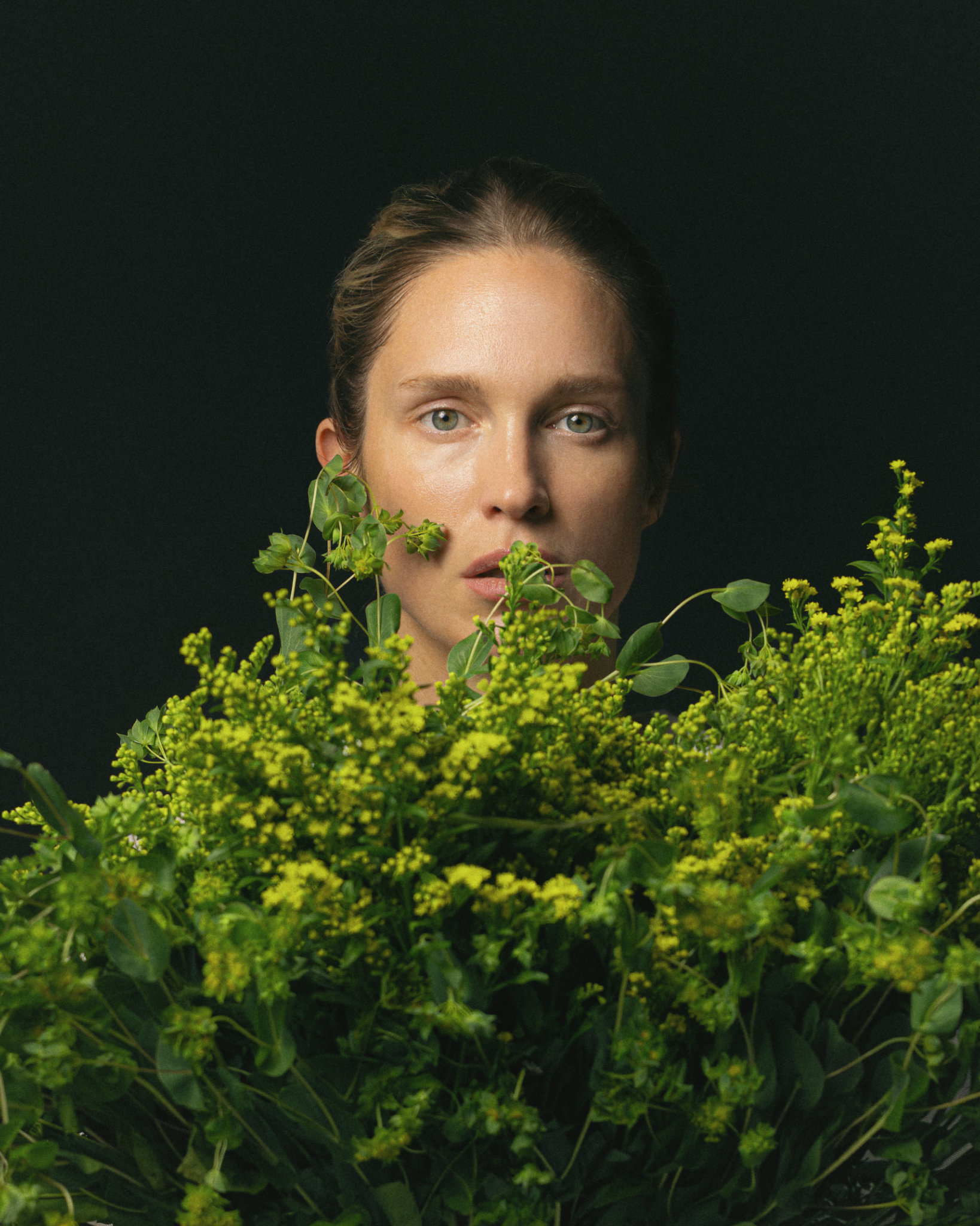 Person peeking out from behind green foliage against a black background